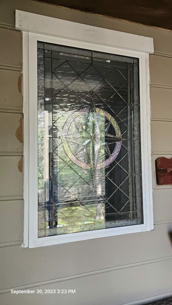 A rectangular, white-framed stained glass window featuring a central starburst design in iridescent glass, surrounded by clear textured glass panels arranged in a grid pattern. The window is set into a light beige exterior wall.