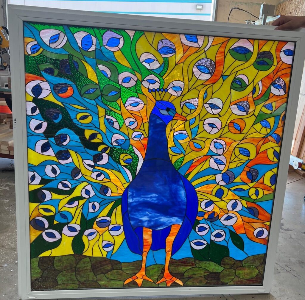 A rectangular stained glass panel in a white frame, depicting a vibrant blue peacock with its tail feathers fanned out. The feathers are composed of colorful blue, green, yellow, and orange glass sections with distinct 'eye' patterns.