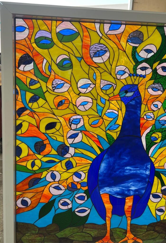 A rectangular stained glass panel depicting a vibrant blue peacock with its tail feathers fully fanned out in yellow, orange, and green glass, featuring numerous blue and white eye-spots.