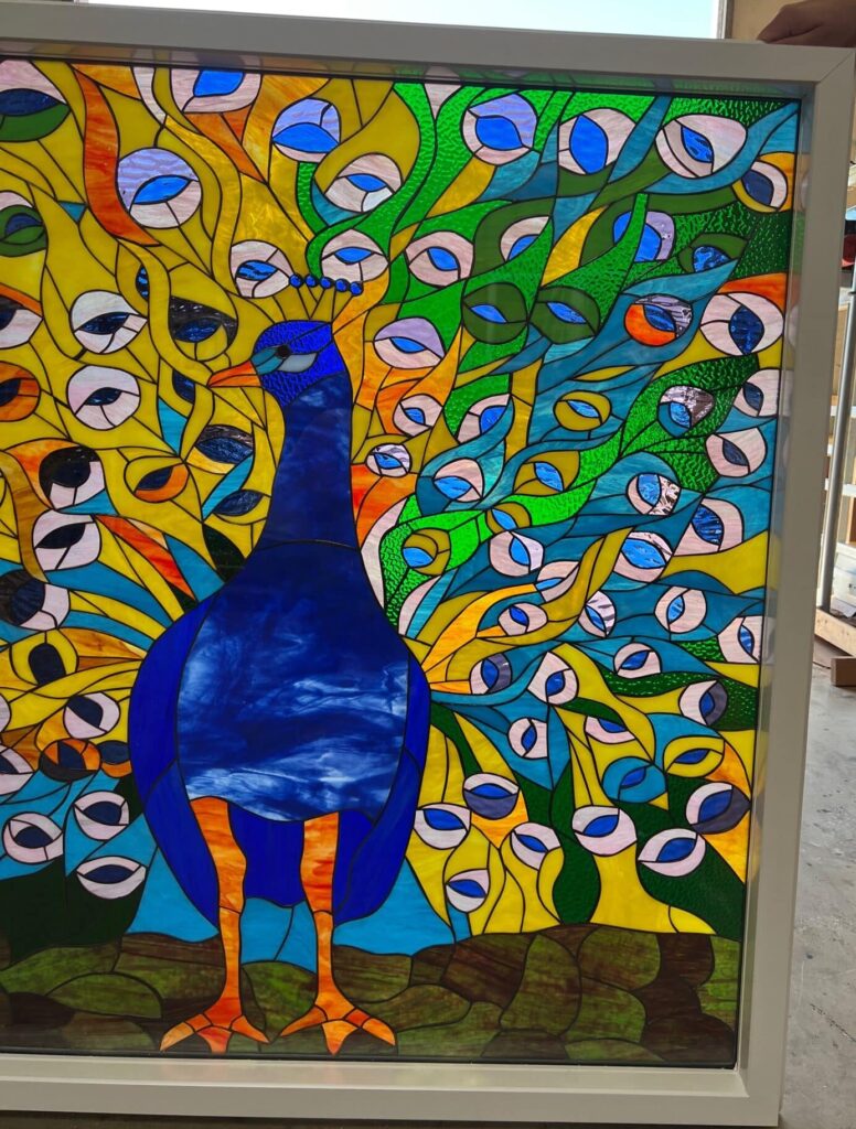 A rectangular stained glass panel in a white frame depicting a full-body peacock. The peacock has a deep blue body and head, with bright orange legs and feet. Its large, fanned tail feathers display a vivid mosaic of yellow, green, and blue glass, featuring numerous white and light blue oval 'eye' patterns.
