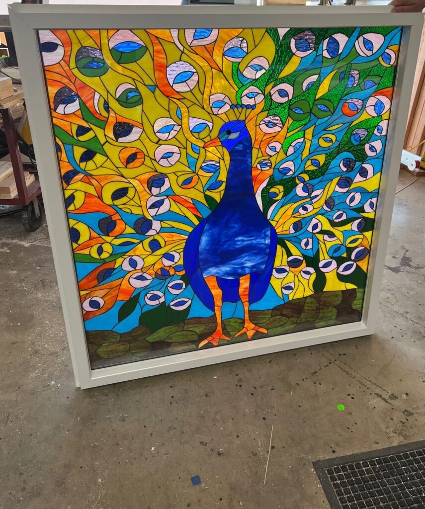 A rectangular stained glass panel featuring a detailed and vibrantly colored peacock with its fanned tail. The peacock's body is deep blue, while its elaborate tail feathers burst with abstract patterns in yellow, orange, green, and sky blue, framed in white.