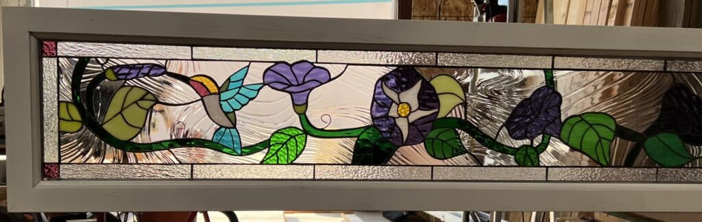 A horizontal stained glass transom panel in a light gray frame, depicting a multicolored hummingbird flying towards purple and yellow morning glory flowers, against a clear textured glass background.