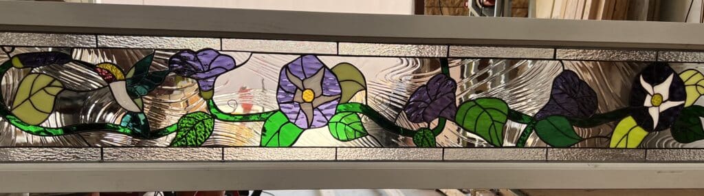A rectangular stained glass transom panel showcasing a floral scene with purple, dark blue, and white morning glory flowers, winding green vines, and lush leaves. A green and yellow hummingbird is depicted on the left. The background consists of clear, textured stained glass, including wavy and hammered patterns, all within a white frame.
