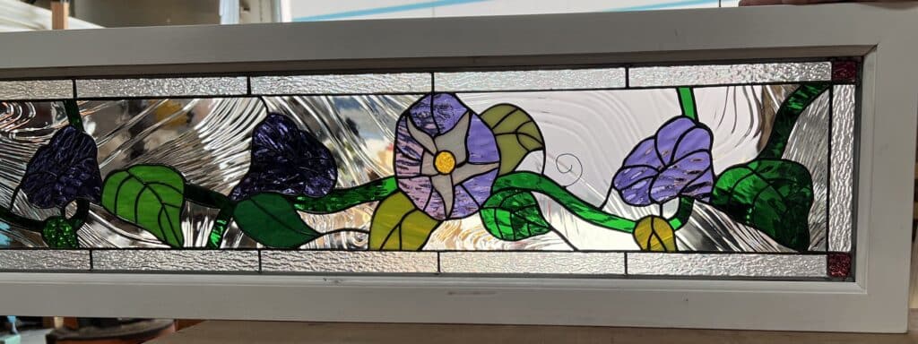 A rectangular stained glass panel depicting purple and lavender morning glory flowers with green vines and leaves, set against clear textured and rippled glass, all framed in white.