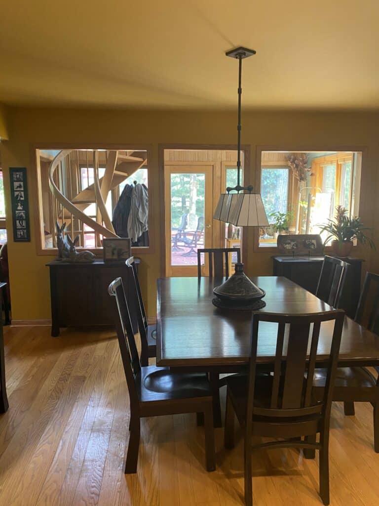A warm and inviting dining room featuring a long wooden table, dark wooden chairs, and a central geometric stained glass chandelier. Three large wooden-framed windows in the background reveal a rustic spiral staircase, outdoor patio, and greenery, offering opportunities for custom stained glass privacy panels.
