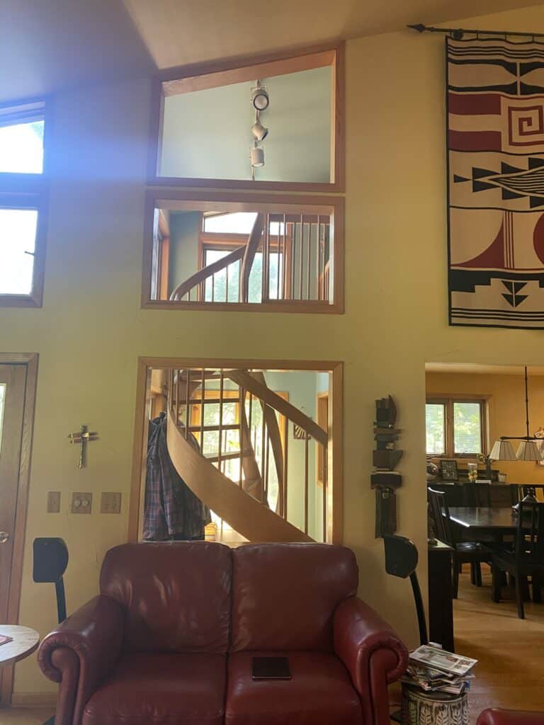 A living room with a red leather couch facing a tall wall featuring three stacked, wooden-framed openings. Through the openings, a curved wooden spiral staircase is visible. This setup is ideal for custom stained glass panels.