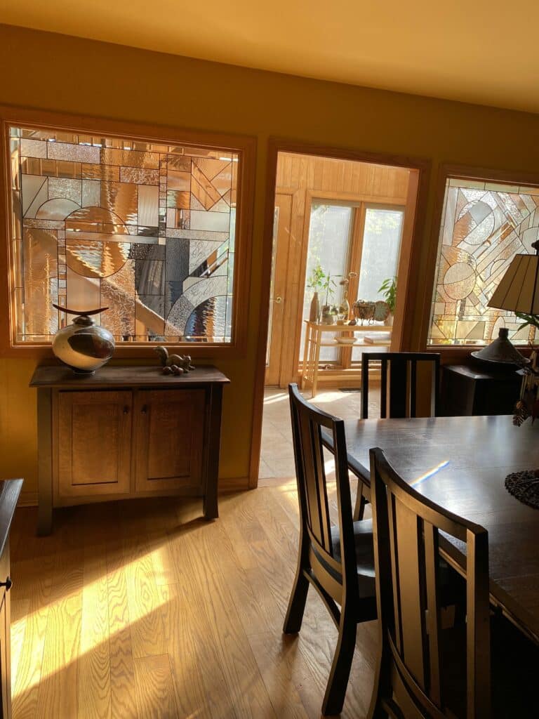 Two large rectangular clear textured stained glass windows with abstract geometric designs, framed in wood, flank a doorway in a sunlit dining room with a dark wood table and chairs.