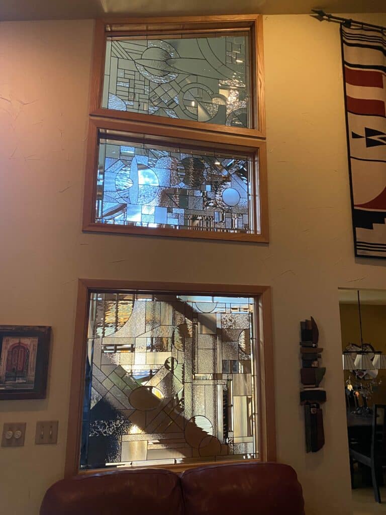 Three vertically stacked rectangular stained glass panels with abstract geometric designs using clear textured and bevelled glass, set in natural wood frames.