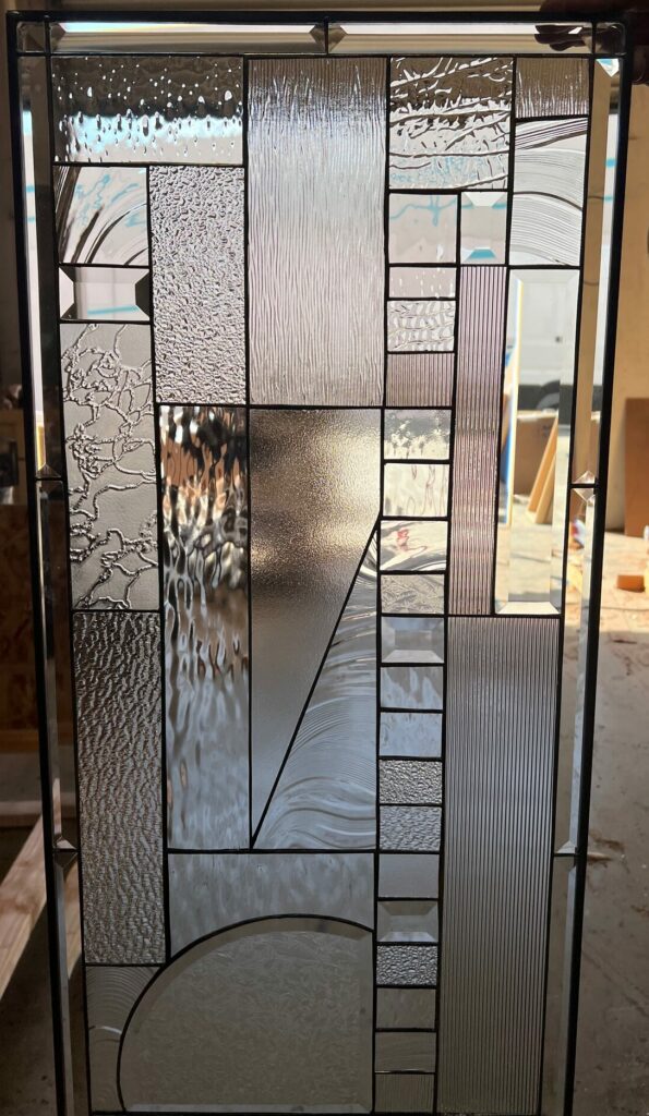 A rectangular, leaded stained glass panel with an abstract geometric design crafted from multiple clear textured glass panels and some clear bevels, enclosed in a dark metal frame.