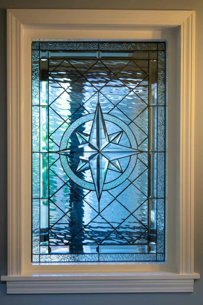 A rectangular stained glass panel featuring a central beveled glass compass rose design within a circle, surrounded by clear textured and beveled glass pieces, set in a white window frame.