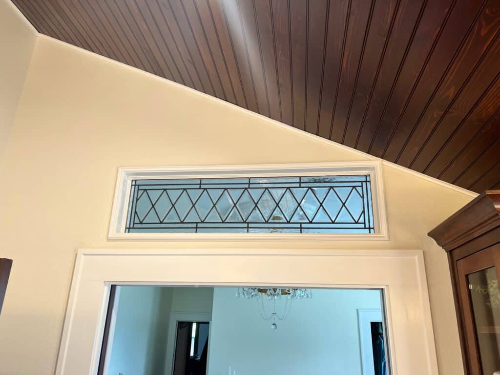 A rectangular clear textured stained glass transom panel featuring a repeating diamond or X-pattern created by black leading, installed above a white-trimmed interior doorway, below a dark wooden plank ceiling.