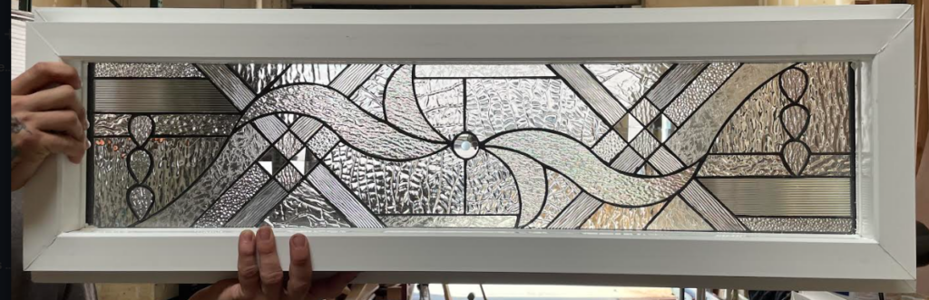 A rectangular stained glass transom panel in a white frame, featuring an abstract design of clear textured glass, including wavy lines, geometric shapes, and some small clear bevels, held by two hands.