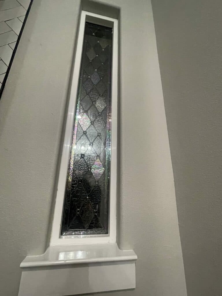 A tall, narrow, rectangular leaded stained glass panel featuring clear textured and beveled diamond-shaped glass pieces, reflecting iridescent light. It is set into a white frame in a light gray wall above a small white sill.