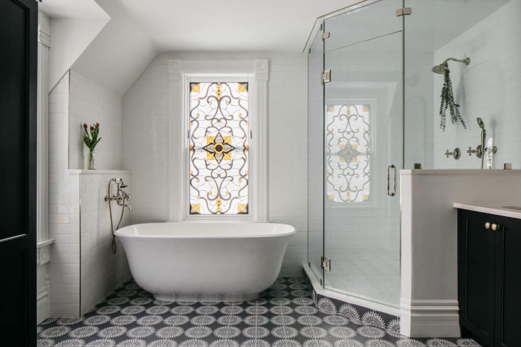 A rectangular stained glass window featuring an intricate Victorian scrollwork pattern with clear, yellow, and dark gray glass, installed above a freestanding white bathtub in a bathroom.