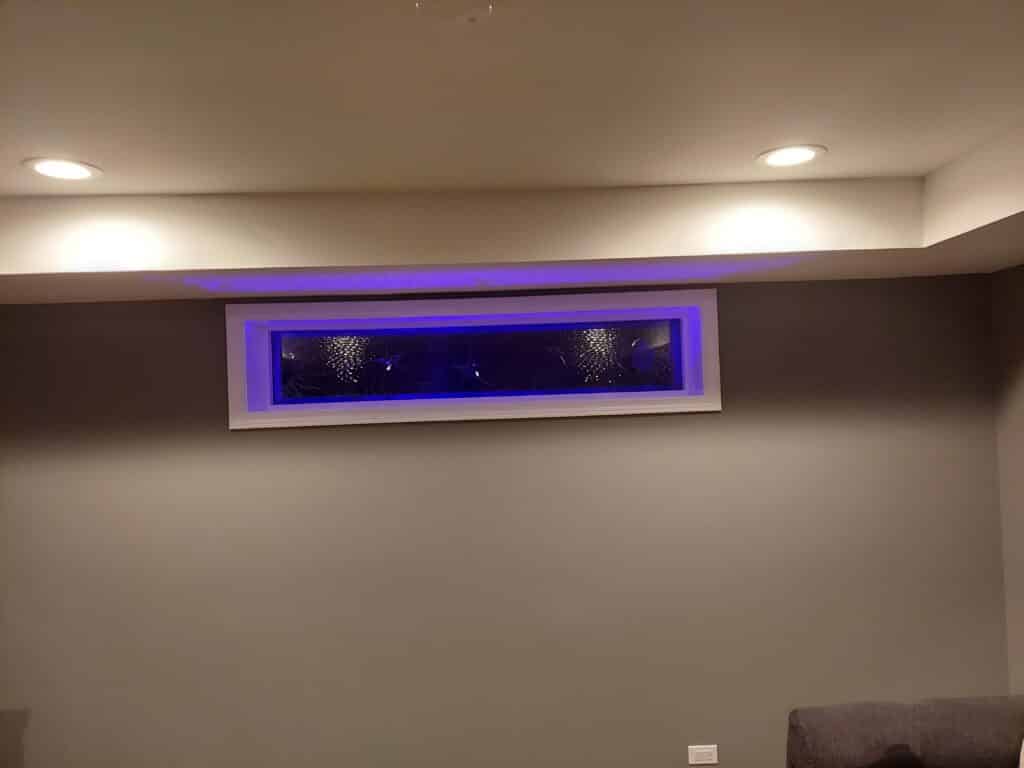A horizontal rectangular decorative privacy glass panel with a white trim, featuring textured glass and illuminated from behind with bright blue LED lighting, set into a gray wall in a modern room.