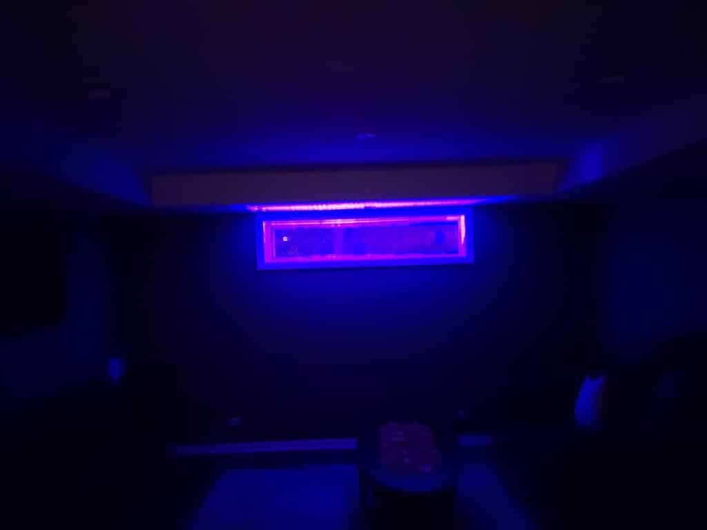 A rectangular stained glass panel in a dark basement window, dramatically illuminated from above by a vibrant purple LED light strip, creating an ethereal glow on the clear, textured glass within the window frame.