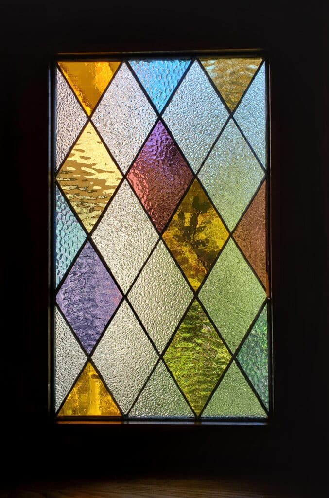 A rectangular leaded stained glass panel featuring a geometric diamond (harlequin) pattern with various textured glass types in clear, yellow, blue, purple, amber, and green colors, framed by dark wood.