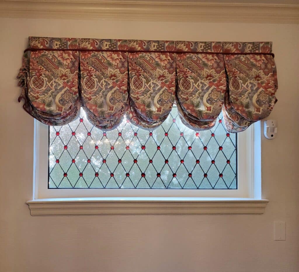Rectangular stained glass window with a diamond pattern of clear textured glass and red circular jewels at each intersection, set in a white frame, partially covered by a floral fabric valance.