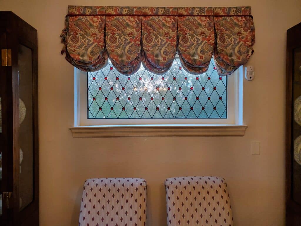 A rectangular leaded stained glass window features a clear, textured diamond lattice pattern, embellished with ruby red glass jewels at each intersection. Sunlight creates a sparkling effect through the glass.