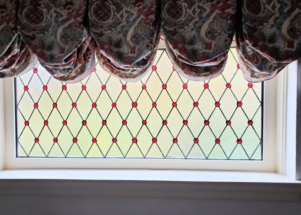 A rectangular stained glass window panel featuring a diamond leaded pattern with clear textured glass. Small, round ruby red glass jewels are set at the intersections of the lead lines. A patterned fabric valance partially covers the top.