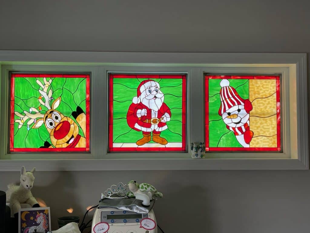 A set of three rectangular stained glass window panels, each with a red border. The left panel features Rudolph the Reindeer with lighted antlers and a red nose on a green textured background. The middle panel shows Santa Claus in his red suit and white beard on a green textured background. The right panel depicts a snowman with a red and white striped hat and scarf, peeking from the side, against a yellow textured background.