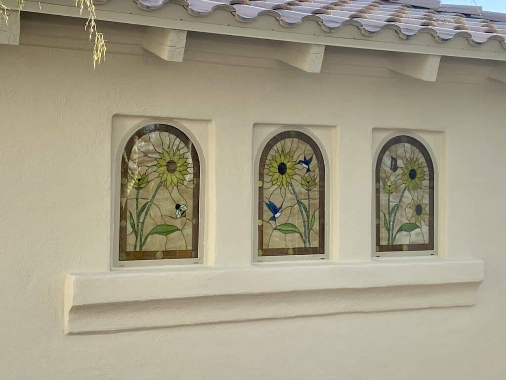 Three arched, insulated stained glass windows featuring vibrant sunflower designs, each with different wildlife (bee, hummingbirds, butterfly), installed in a cream stucco wall beneath a terra cotta tiled roof.
