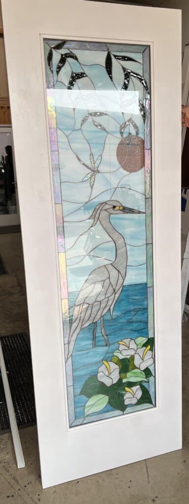 A tall, rectangular stained glass door panel depicting a gray heron standing in blue water, with white calla lilies, green leaves, a dragonfly, and a brown sun-like circle under a pale blue sky. The panel features iridescent glass accents.