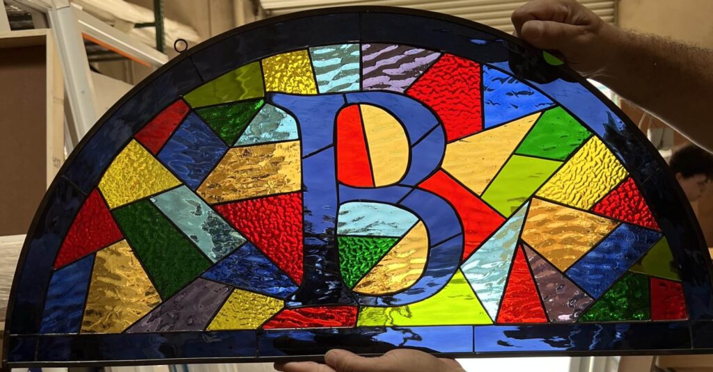An arched, leaded stained glass panel featuring a bold blue Bitcoin 'B' symbol surrounded by vibrant, textured multi-colored geometric glass pieces.