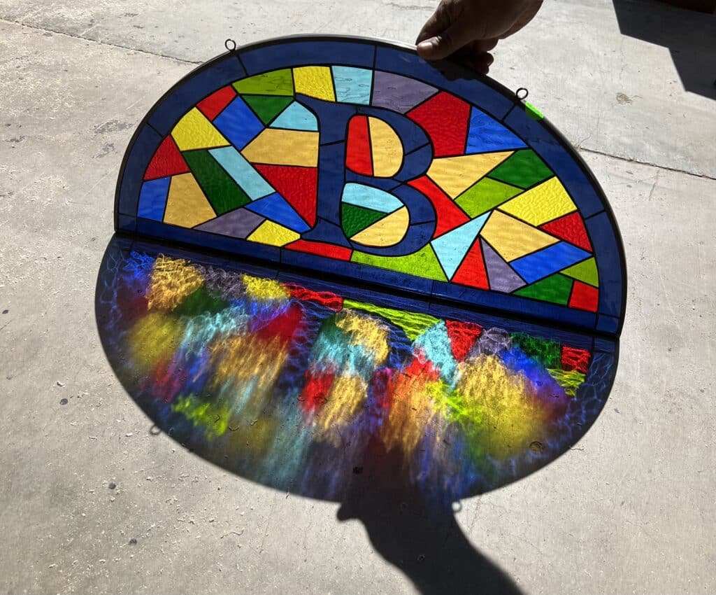 A person's hand holds up a vibrant semicircular leaded stained glass panel featuring a large letter 'B' in its center. The panel is bordered in deep blue, with the 'B' and surrounding abstract geometric shapes made from various textured colored glass, including red, yellow, green, blue, purple, and light blue. The panel casts dynamic, colorful light patterns onto the concrete ground below.