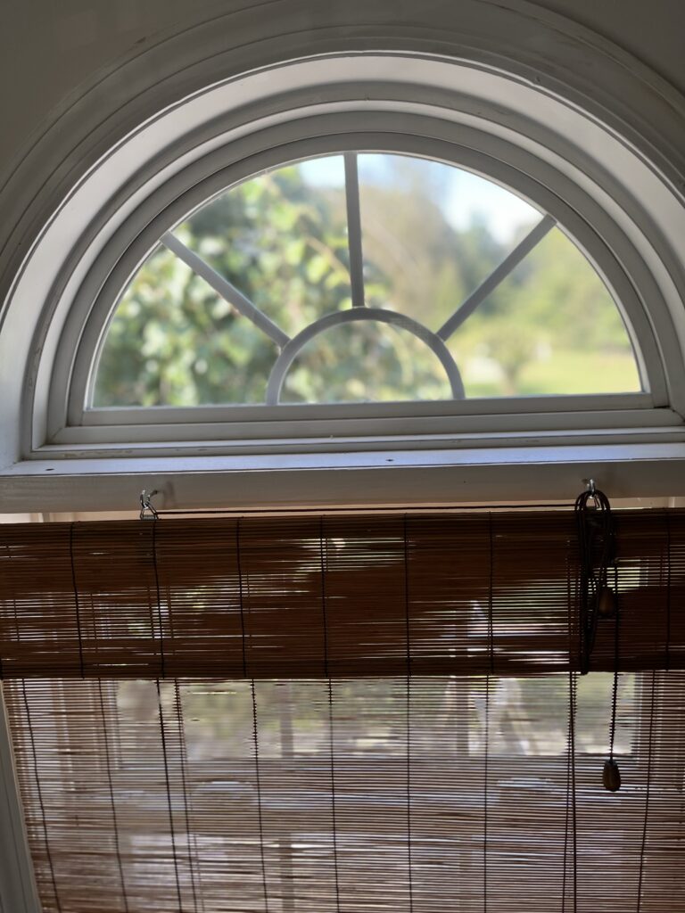 An arched transom window with a decorative white grille and a partially lowered bamboo shade, revealing a blurred green outdoor scene.