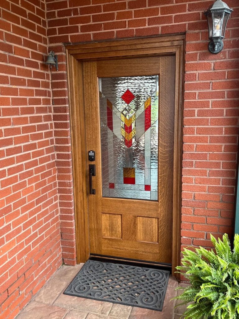 A wooden front door featuring a rectangular Arts and Crafts style stained glass panel with geometric clear, red, and amber-colored glass, set into a red brick wall.