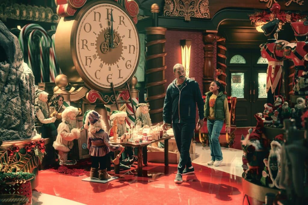An adult man and a young girl stroll through a fantastical Christmas workshop with a giant golden clock, huge candy canes, and numerous elf-like dolls on a shiny red floor.