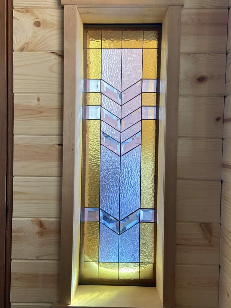 A rectangular stained glass panel featuring a central geometric chevron pattern in clear beveled glass, flanked by vertical sections of amber and clear textured glass. The panel is set within a light wooden frame and surrounded by a light wood-paneled wall.