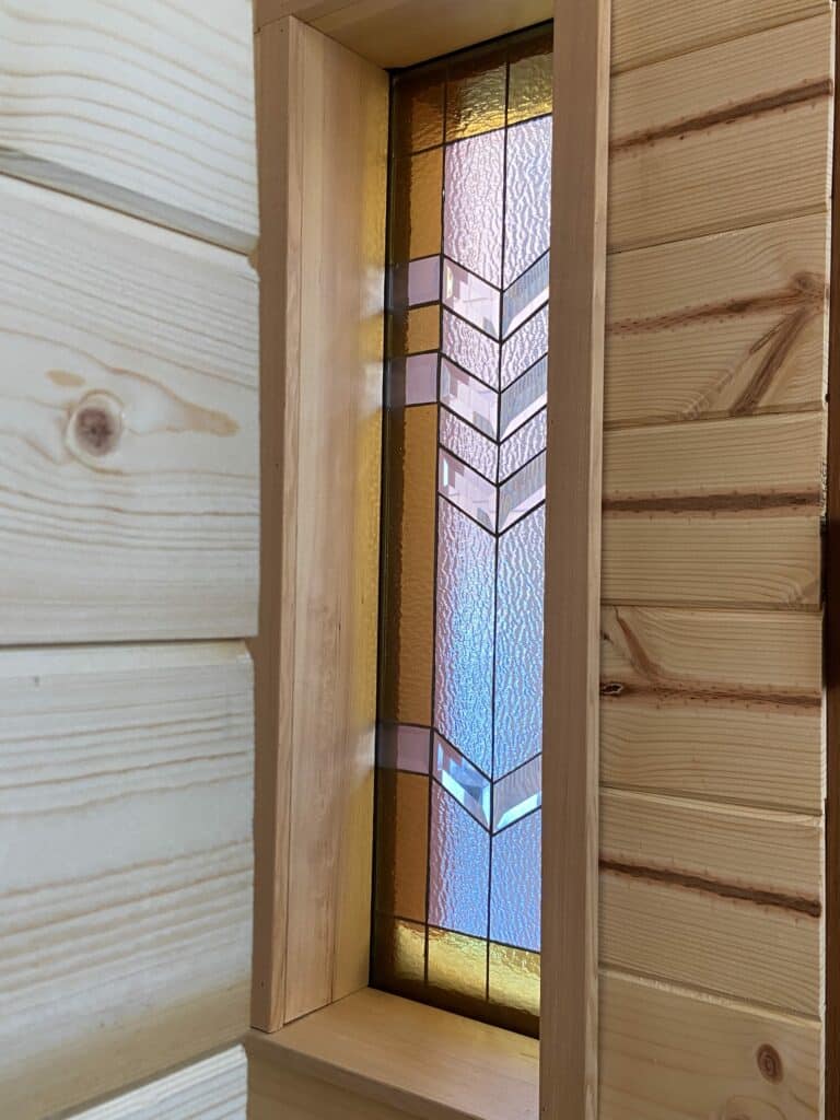 A rectangular stained glass window panel featuring a central chevron pattern of clear textured glass and bevels, flanked by amber glass, with small lavender accent pieces, installed in a light wood frame.