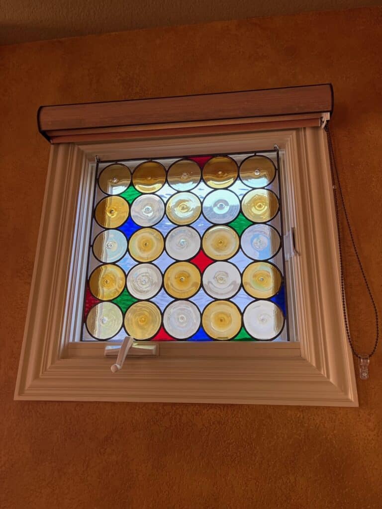 A square stained glass panel installed in a window, featuring a grid of multi-colored circular rondel glass pieces. The colors include amber, clear, blue, green, and red, with visible swirls in each rondel. The panel is set within a white window frame, under a light wood window blind, against a textured orange wall.