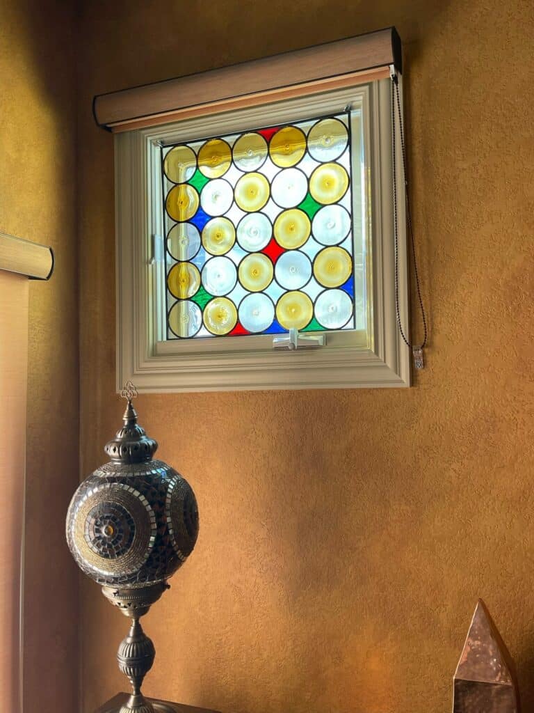 A rectangular stained glass window with a grid pattern of clear, yellow, blue, red, and green circular bullseye glass pieces.
