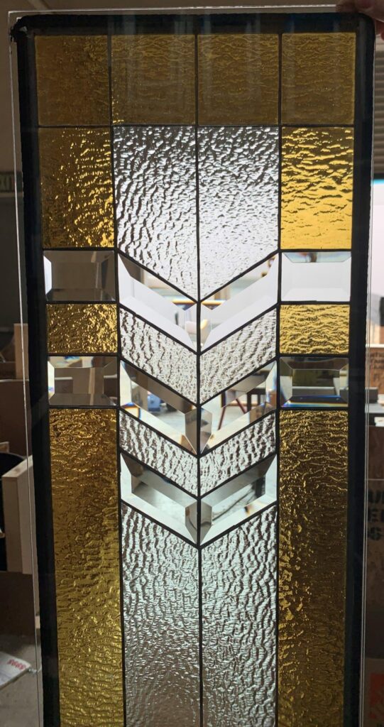 A rectangular leaded stained glass panel with an Art Deco geometric design. It features a central vertical chevron pattern composed of clear textured glass and clear bevels, surrounded by textured gold glass and small clear rectangular bevels, all joined by dark lead lines.