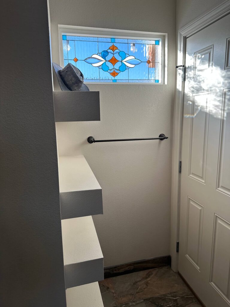 A rectangular stained glass window featuring a symmetrical, geometric design with blue, orange, and white glass, set into a white wall.