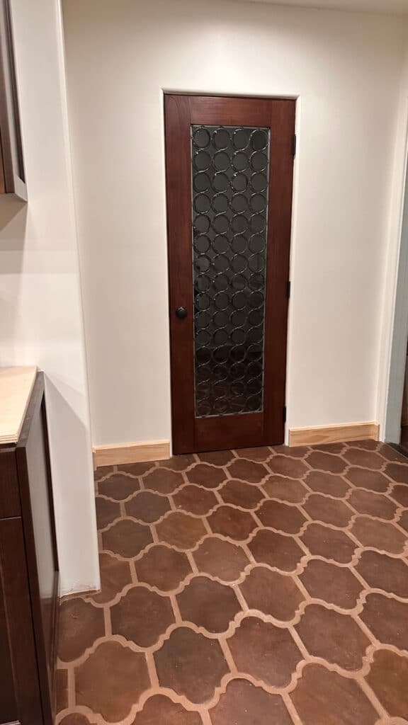 A dark wood interior door featuring a full-length stained glass panel composed of clear, circular rondel glass pieces, installed in a light-colored wall above a terracotta arabesque tile floor.