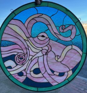 A circular stained glass panel featuring a large pink and yellow octopus with swirling tentacles, surrounded by various shades of blue textured water, all framed by a dark green border.