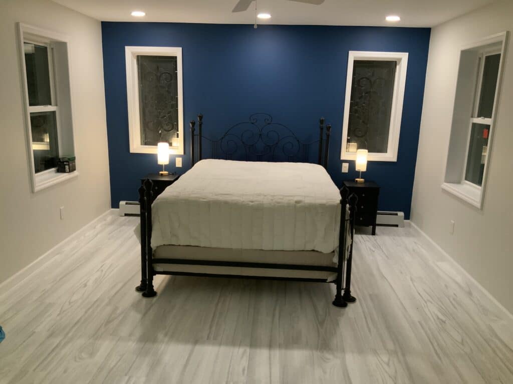 A bedroom with a dark blue accent wall, featuring a black wrought iron bed with white textured bedding, and two rectangular windows fitted with dark, ornate privacy glass inserts. Two black nightstands with modern cylindrical lamps flank the bed, and the room has light gray wood-look laminate flooring.