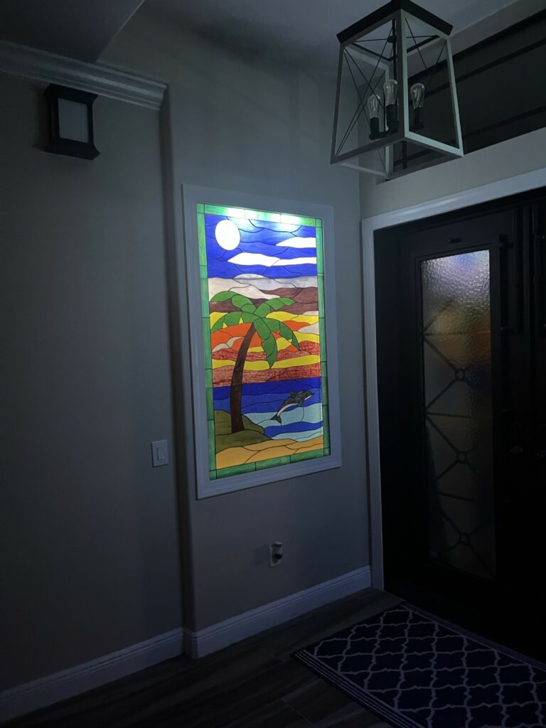 A rectangular stained glass panel depicting a vibrant tropical scene with a palm tree, a dolphin jumping from water, and a prominent moon against a layered blue and orange sky.