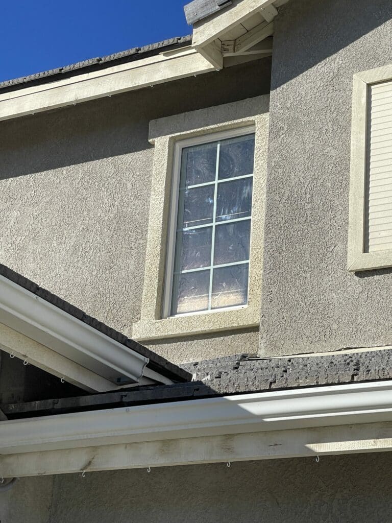 A rectangular window with white grid lines (muntins) in a light beige stucco wall. A white rain gutter and dark roof edge are visible below the window against a blue sky.