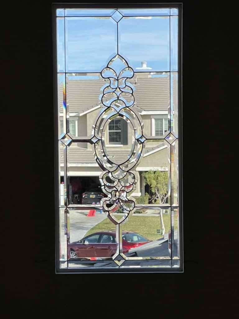 A rectangular clear beveled stained glass panel viewed from inside, showing an elaborate, symmetrical central scrollwork design composed of numerous curved and straight beveled glass pieces, all framed by larger clear rectangular bevels. The leading is dark. The panel is set in a window looking out onto a suburban street with a house, driveway, and a red car.