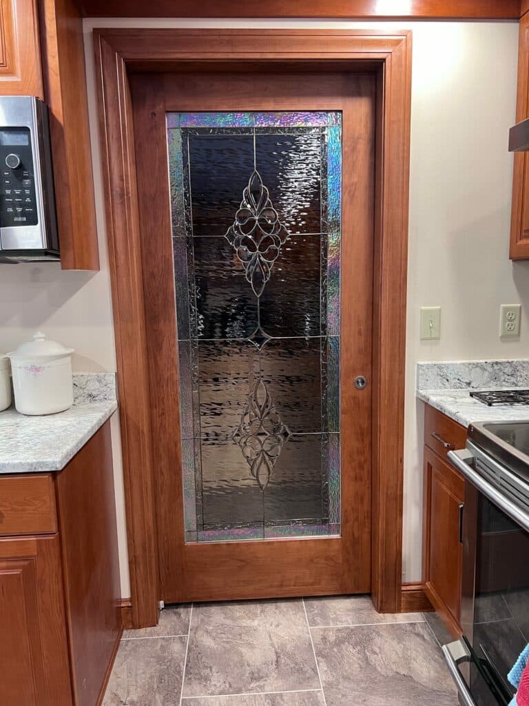 A full-height, cherry wood pantry door with an integrated stained glass panel. The panel features clear textured glass, two ornate beveled scrollwork medallions, and an iridescent rainbow-hued border, installed in a modern kitchen with granite countertops.