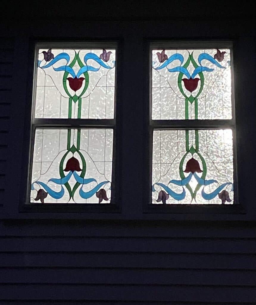 Two rectangular stained glass windows, each composed of two panels, featuring a symmetrical Art Nouveau design. The pattern includes red bell flowers, green stems and leaves, flowing blue ribbon-like elements, and small purple buds, all set against a textured clear glass background with leaded lines, seen from outside at night.