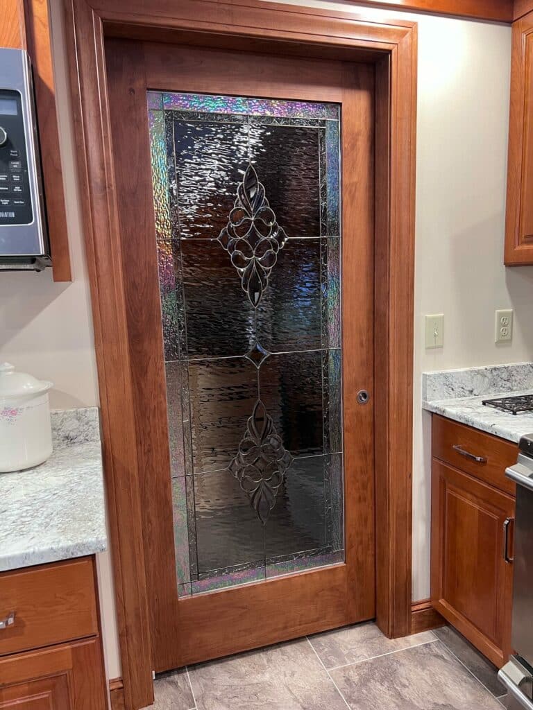 A full-view wooden pantry door with a clear textured and iridescent stained glass panel, featuring a central leaded floral and scroll design, set in a modern kitchen.
