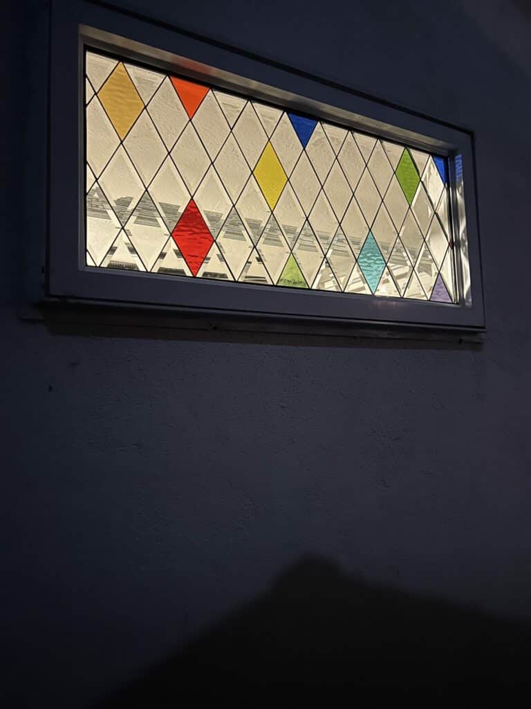 A rectangular stained glass window with a diamond grid of clear textured glass, interspersed with colorful rhombus pieces in red, yellow, blue, green, aqua, orange, and violet, set in a white frame against a dark wall and illuminated from behind.