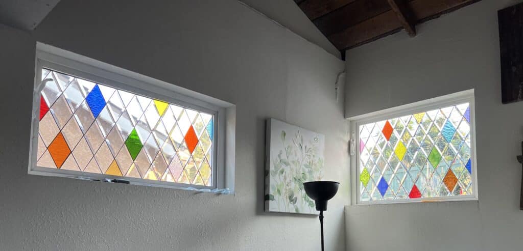 Two rectangular transom windows with a colorful harlequin stained glass design, featuring clear textured diamond panes and scattered colored diamonds in red, orange, yellow, green, and blue.