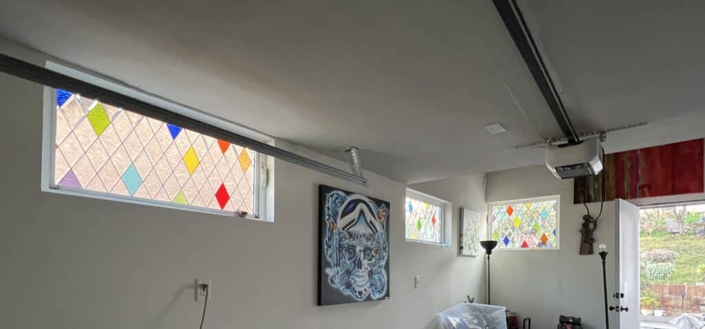 A series of rectangular stained glass panels, each featuring a clear diamond grid with randomly placed colored diamond accents in blue, green, yellow, orange, and red. They are installed in a room with a garage door track visible.